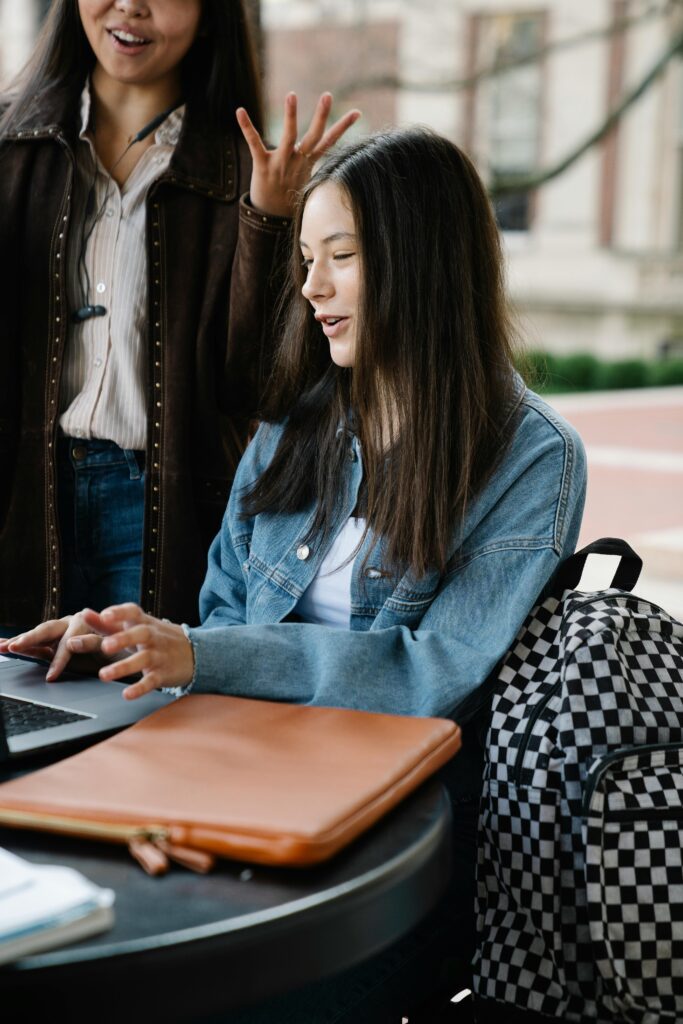 Two young women collaborating on a laptop at an outdoor table.